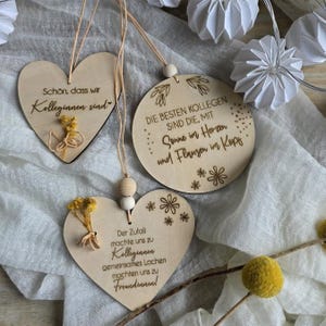 Puede incluir: Adornos de madera en forma de corazón y circulares con texto grabado y detalles florales. Los adornos en forma de corazón tienen flores secas adjuntas. El texto incluye frases como "colegas" y "amigos". Los adornos están suspendidos por hilo.
