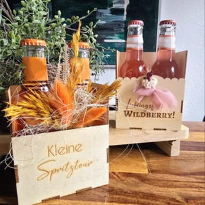 Könnte beinhalten: Zwei Holzkisten, eine mit orangefarbenen Flaschen und Trockenblumen, die andere mit rosafarbenen Flaschen. Die erste Kiste trägt den Text "Kleine Spritztour". Die zweite Kiste trägt den Text "Lieblings Wildberry!"