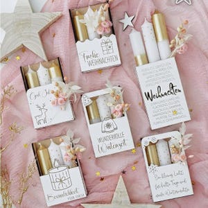 Könnte beinhalten: Sechs rechteckige weiße Geschenkboxen mit Kerzen, Trockenblumen und winterlichen Texten. Jede Box enthält goldene und weiße Kerzen, kleine Blumenarrangements sowie schwarze Grafiken wie ein Rentier, einen Pullover und ein Geschenk.