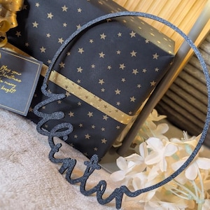 Op de afbeelding: Een donkergrijze glitter "Winter" woordkunst decoratie in een cirkelvorm. De woordkunst staat voor een ingepakt cadeau met een zwarte achtergrond en gouden sterren. Een cadeaulabel luidt "May your Christmas be merry & bright!"