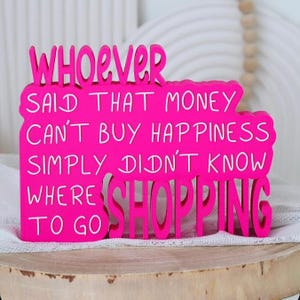 Peut inclure: Un panneau en bois rose vif avec des lettres blanches indiquant : WHOEVER SAID THAT MONEY CAN'T BUY HAPPINESS SIMPLY DIDN'T KNOW WHERE TO GO SHOPPING. Le panneau présente un style découpé audacieux avec une apparence superposée et des bords lisses.