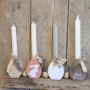 Może przedstawiać: Cztery wielkanocne świeczniki z białymi świecami. Każdy uchwyt w kształcie jajka jest ozdobiony motywem królika i napisami "Frohe Ostern" lub "Kusiner Ostergruss". Uchwyty znajdują się na drewnianej półce.