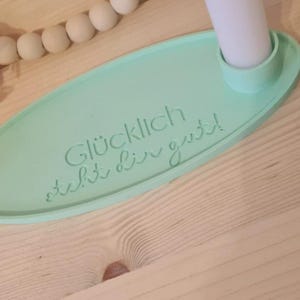 May include: A light green oval-shaped candle holder with the text "Gl&uuml;cklich steht dir gut!"  The holder has a small indentation for a candle.