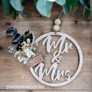 May include: A wooden Mr. & Mrs. sign with a floral arrangement and a small tag that says "Herzlichen Glückwunsch". The sign is round and has a natural wood finish. The floral arrangement includes white and cream-colored flowers and greenery.