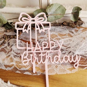 May include: A light pink cake topper with the words "Happy Birthday" in a cursive font. The topper features a gift box design with a bow on top, perfect for a birthday celebration.