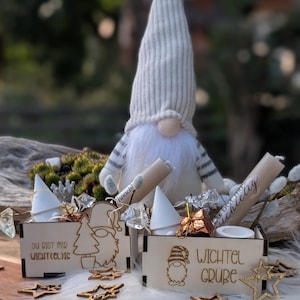 Könnte beinhalten: Eine festliche Wichtelpuppe mit langem weißem Bart und gestreifter Mütze sitzt hinter zwei Holzkisten. Die Kisten sind mit Laserschnitten von Wichteln, Bäumen und Sternen verziert und enthalten kleine Geschenke. Der Text "DU BIST MIR WICHT(EL)IG" und "WICHTEL GRUBE" ist sichtbar.