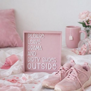 May include: A pink framed sign with the text "PLEASE LEAVE DRAMA AND DIRTY SHOES OUTSIDE" is next to a pair of pink sneakers. A pink mug with a tea bag, a spilled bottle of nail polish, and a jar of pink candies are also in the image.