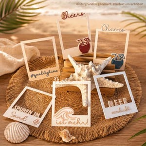 Può includere: Una collezione di sei cornici portafoto rettangolari bianche con vari accenti decorativi e testi tra cui Cheers, #qualitytime, Meer, SPUREN IM Sand, Meer ist mehr! e MEER verliebt, disposte attorno a una stella marina bianca centrale.