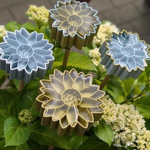 May include: Four decorative sunflower garden stakes in shades of blue and gold. The flower-shaped stakes have a detailed petal design and are mounted on wooden sticks, set against a backdrop of green foliage and white flowers.