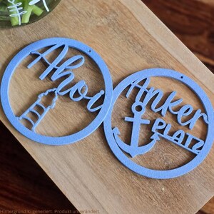 May include: Two circular light blue decorative cutouts. The left cutout features a lighthouse silhouette and the text Ahoi. The right cutout features an anchor silhouette and the text Anker PLATZ. Both items have a small hole at the top for hanging.