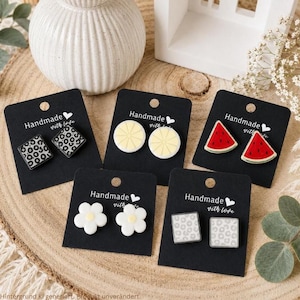 May include: Five pairs of stud earrings displayed on black cards labeled Handmade with love. Designs include black and white leopard print squares, yellow lemon slices, red watermelon wedges, white flowers with yellow centers, and light gray leopard print squares.