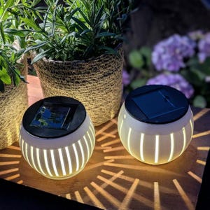 May include: Two solar-powered garden lights with a cream-colored, spherical design and vertical cutouts. Each light has a black top with a solar panel. The lights are on a wooden surface, with plants in woven baskets in the background.