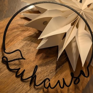 Puede incluir: Una corona de metal negro con la palabra "Zuhause" en escritura cursiva, que encierra una decoración de papel en forma de estrella de color crema. La estrella está hecha de papel plegado y se encuentra sobre una superficie de madera.