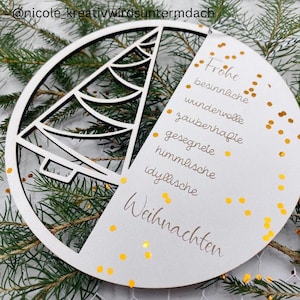 Könnte beinhalten: Weihnachtsornament aus weißem Holz mit einem ausgeschnittenen Weihnachtsbaumdesign in einem Kreis. Das Ornament hat einen Abschnitt mit den deutschen Worten "Frohe Weihnachten" und anderen festlichen Wörtern. Goldener Glitzer akzentuiert das Design.