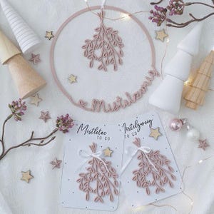 May include: A festive holiday scene featuring a pink mistletoe wreath and matching mistletoe decorations. The wreath has the word "Mistletoe" in cursive. Two cards with mistletoe designs and the words "Mistletoe to go" are also present, along with wooden stars and decorative trees.