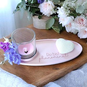 May include: A pink oval tray with the text "Du bist ph&auml;nomenal!"  A white heart-shaped object and a glass candle holder with a pink candle are on the tray.