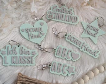 Könnte beinhalten: Ein Set aus sieben pastellgrünen Schlüsselanhängern mit verschiedenen Designs und Texten. Die Schlüsselanhänger zeigen die Wörter "School Kid", "I am in 1st Grade", "ABC" und "Endlich Schulkind".