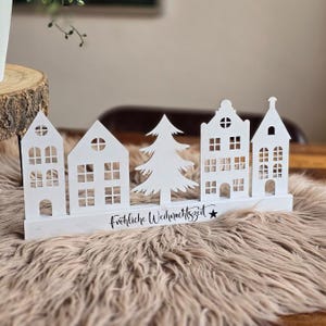 Può includere: Decorazione natalizia in legno bianco con case stilizzate e un albero di Natale. La scritta "Fröhliche Weihnachtszeit" è presente. L'ornamento è posto su una superficie soffice di colore marrone chiaro.