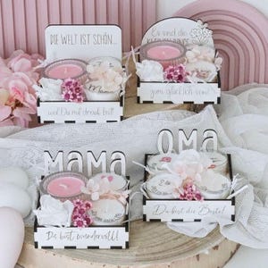 May include: Four decorative gift boxes with candles, flowers, and text. Each box is white with black accents and contains a pink candle, white flowers, and decorative elements. The boxes have German text, including "Mama" and "Oma."