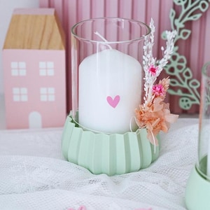 Può includere: Una candela bianca con un design a forma di cuore rosa si trova in un portacandele di vetro verde con una base decorativa. Fiori secchi sono disposti intorno al portacandele.