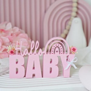Puede incluir: Letras de madera rosa que deletrean "Hallo Baby" con un recorte de arcoíris rosa detrás de la "B". Las letras están sobre una superficie blanca con un fondo a rayas rosa y blanco.