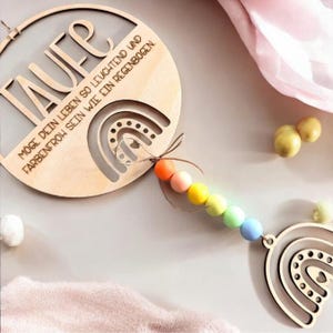 Może przedstawiać: Drewniana dekoracja w tęczowym motywie ze słowem "TAUFE" i niemieckim tekstem. Projekt obejmuje wycięcie w kształcie tęczy i sznur kolorowych koralików. Dekoracja wykonana jest z jasnego drewna.