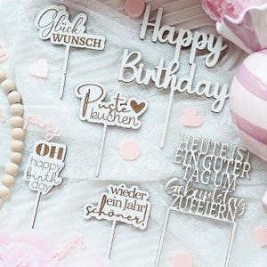 Könnte beinhalten: Eine Sammlung von Kuchenaufsätzen aus Holz mit verschiedenen Schriftzügen, darunter "Happy Birthday" und deutsche Sätze. Die Aufsätze sind in einer Schreibschrift gehalten und sind weiß und braun. Rosa herzförmige Konfetti sind verstreut.