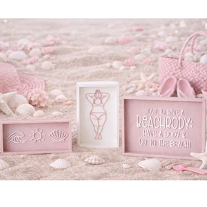 May include: Pink beach-themed decor on sand. Includes framed art with wave, sun, and seashell designs, a framed silhouette of a person in a swimsuit, and a sign that reads "How to have a beach body: Have a body & go to the beach!"