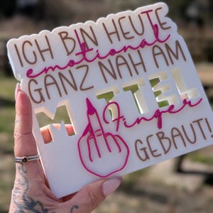 May include: A white sign with brown and pink text that reads "ICH BIN HEUTE emotional GANZ NAH AM MITTEL Finger GEBAUT!" The sign features a cutout middle finger graphic in pink.