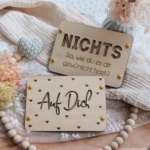 May include: Two wooden signs with gold accents. The top sign says "Nichts So, wie du es dir gew&uuml;nscht hast:" and the bottom sign says "Auf Dich" with small hearts surrounding the text.