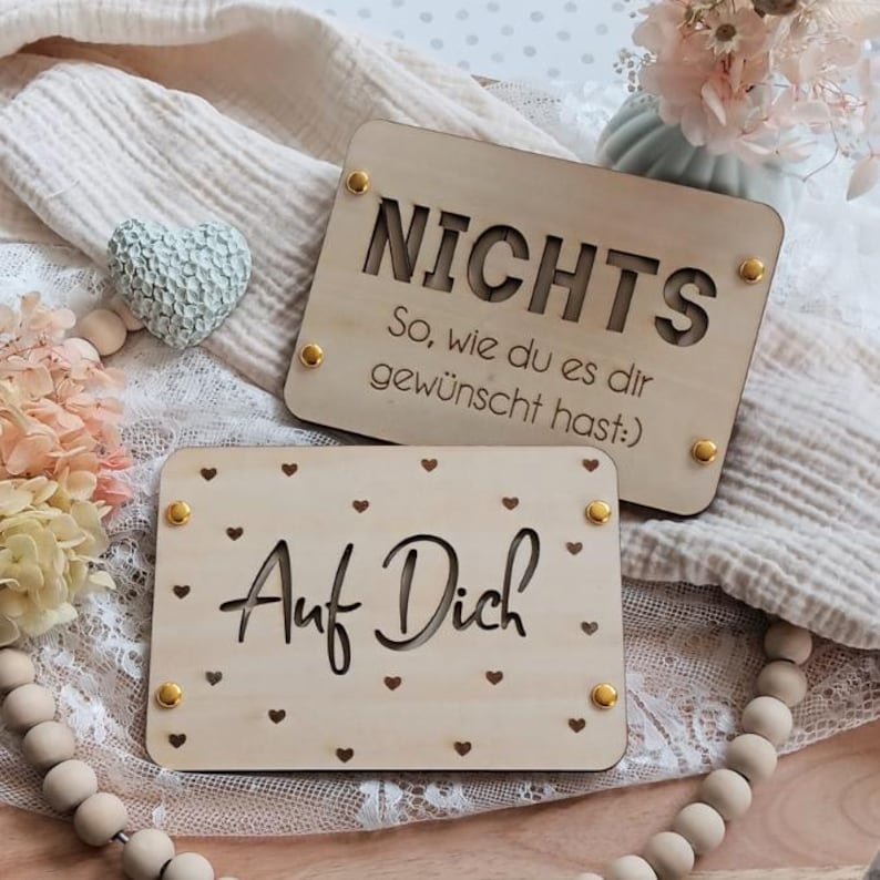 May include: Two wooden signs with gold accents. The top sign says "Nichts So, wie du es dir gew&uuml;nscht hast:" and the bottom sign says "Auf Dich" with small hearts surrounding the text.