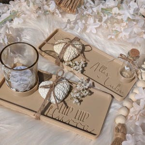 Puede incluir: Dos bandejas decorativas con el texto "Hip Hip Hooray" y "Alles Liebe" en ellas. Cada bandeja tiene una pequeña decoración en forma de corazón blanco con un lazo de cuerda marrón, y un pequeño frasco de vidrio con tapa de corcho. Una bandeja también tiene un pequeño portavelas de vidrio.