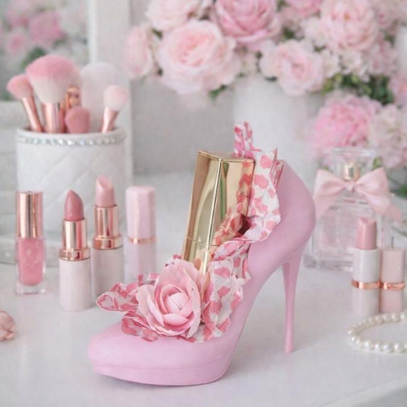 May include: A pink high-heeled shoe decorated with a pink flower and a gold lipstick tube. The shoe is surrounded by pink lipsticks, makeup brushes, and a perfume bottle, all in a soft, feminine aesthetic.