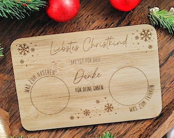 Op de afbeelding: Een rechthoekige houten serveerplank met gegraveerde tekst en twee cirkelvormige uitsparingen. De tekst bevat "Liebstes Christkind" en "Danke". Decoratieve sneeuwvlokken zijn ook gegraveerd. Rode kerstballen en groen in de achtergrond.