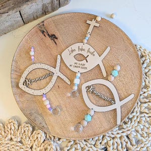 May include: Wooden fish-shaped ornaments with the word "Kommunion" and other text. Each ornament is adorned with beads in pastel colors and a clear crystal. The ornaments are displayed on a wooden platter.
