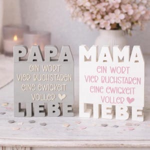 以下が含まれることがあります： 「PAPA」と「MAMA」の文字が大きく書かれた、グレーと白の2つの装飾用木製ブロック。各ブロックにはドイツ語のテキストが追加されており、「LIEBE」（愛）という単語も含まれています。背景には花瓶に入った花があります。