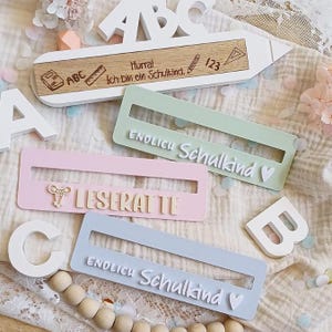 Puede incluir: Letrero de madera con forma de lápiz con el texto "Hurra! Ich bin ein Schulkind." y elementos escolares. Tres letreros rectangulares en colores pastel con los textos "ENDLICH Schulkind" y "LESERATTE".