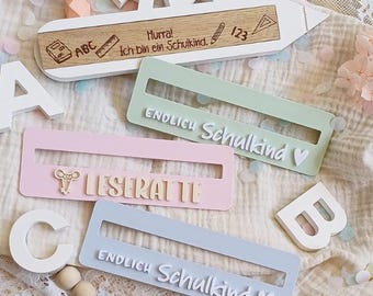Könnte beinhalten: Holzschild in Bleistiftform mit dem Text "Hurra! Ich bin ein Schulkind." und Schulelementen. Drei rechteckige Schilder in Pastellfarben mit den Texten "ENDLICH Schulkind" und "LESERATTE".