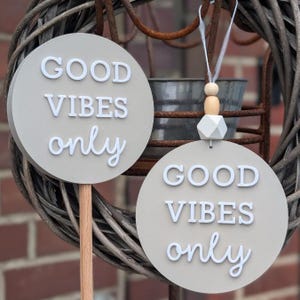 Puede incluir: Dos letreros redondos de color gris claro con las palabras "GOOD VIBES only" en escritura blanca. Un letrero está sobre un palo de madera, el otro cuelga de una cuenta decorativa y una cinta. Los letreros están colocados contra una corona tejida.