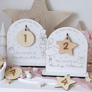 Puede incluir: Calendario de adviento navideño de madera blanca con una estrella y un adorno redondo. El calendario presenta los números 1, 2, 4 y 7, junto con el texto "Wir müssen nur noch" e "Ich muss nur noch". El calendario tiene ilustraciones de un cascanueces y una bailarina.