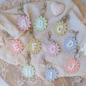 Könnte beinhalten: Eine Sammlung von schlüsselanhängern in Form von Gänseblümchen in verschiedenen Pastellfarben, die jeweils einen Buchstaben des Alphabets tragen. Die Schlüsselanhänger haben silberfarbene Verschlüsse und liegen auf einem cremefarbenen Stoff.