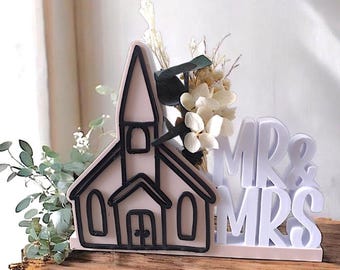 Op de afbeelding: Een taarttopper voor een bruiloft met een kerk silhouet met zwarte contouren en de woorden "MR & MRS" in het wit. De topper is versierd met gedroogde bloemen en groen, geplaatst op een houten oppervlak.
