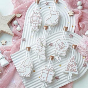 Puede incluir: Viales de vidrio transparente con tapones de corcho, cada uno adornado con un dije navideño blanco y rosa. Los dijes incluyen un cascanueces, un calcetín, un bastón de caramelo, un globo de nieve, un gnomo, una caja de regalo y un árbol de Navidad.