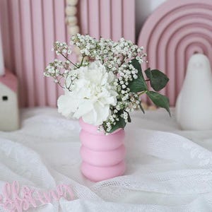 May include: A pink ceramic vase with a textured, rounded design holds a bouquet of white and light pink flowers. The vase is sitting on a white lace tablecloth.