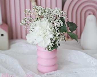 May include: A pink ceramic vase with a textured, rounded design holds a bouquet of white and light pink flowers. The vase is sitting on a white lace tablecloth.