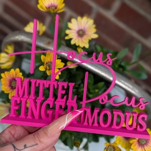 May include: Pink 3D printed sign with the words "Hocus Focus Mittel Finger Modus" in a playful font. The sign is sitting on a table with a hand holding a nail with a white design.