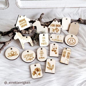 May include: A collection of wooden tags with various designs and sayings, including "Home Sweet Home", "Für Dich", "Das Leben ist schön", and "Heimat Liebe".