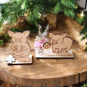 May include: Two wooden pig figurines, one holding a horseshoe and the other a four-leaf clover. A small glass vase with pink flowers and the text "Viel Glück in neuen Jahr" are also present. The figurines are on a wooden surface.