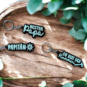 May include: Three black keychains with white and light blue text. The first keychain says "Best Papa" in a cursive font. The second keychain says "Papitan" with a small steering wheel icon. The third keychain says "Du bist der Hammer" with a small hammer icon.