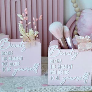 Peut inclure: Deux porte-objets roses avec le texte "Beauty Begins The Moment You Decide To Be Yourself". L'un contient des pinceaux de maquillage, l'autre des fleurs séchées. Les supports sont sur une surface en dentelle.